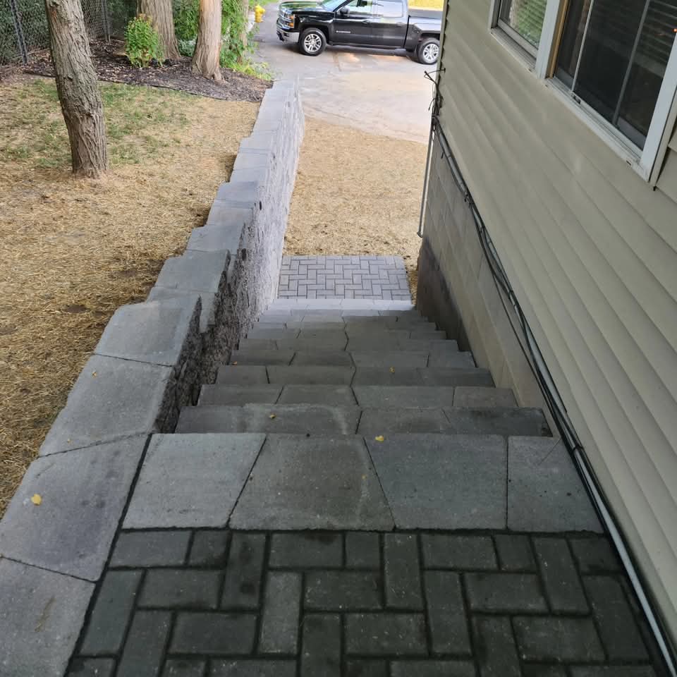 Raised herringbone-pattern paver landing with block steps leading up next to an AC unit and house siding