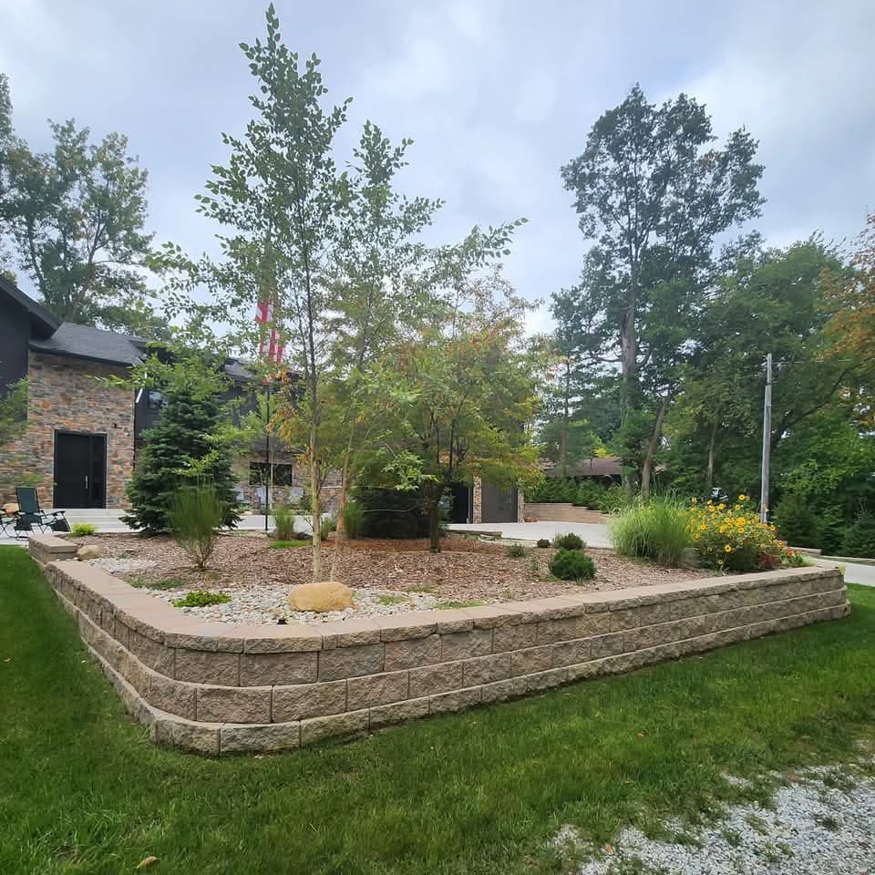 Raised tan block planter with young birch trees and river rock in front of a stone-faced home