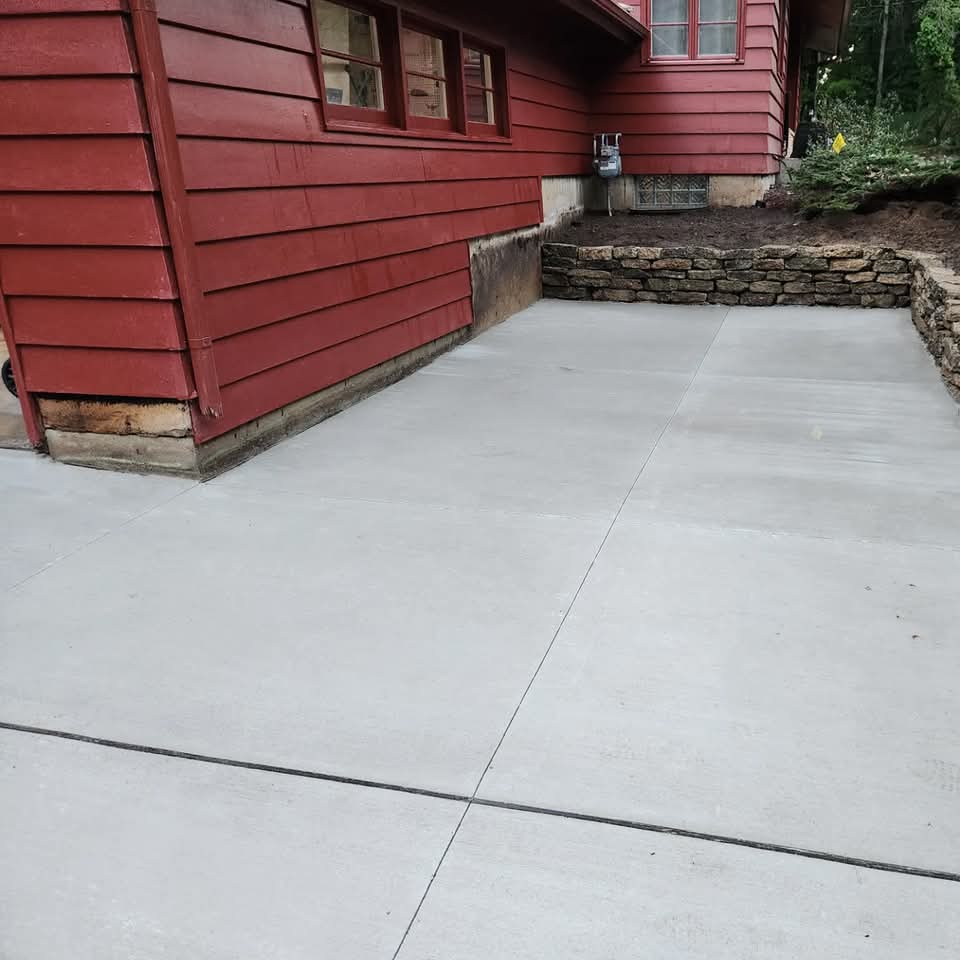 Freshly poured concrete pad next to a red house with a natural stone retaining wall and wooded hillside