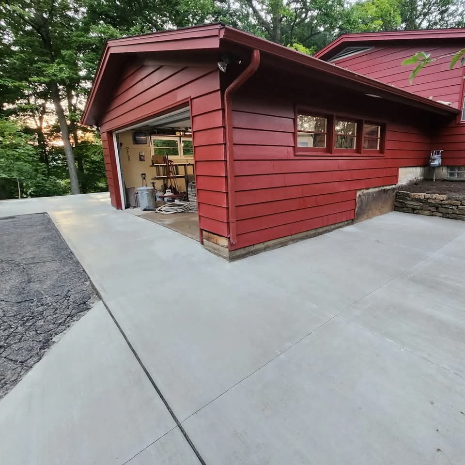 New concrete driveway and pad wrapping around a red house with open garage and old cracked asphalt at the edge