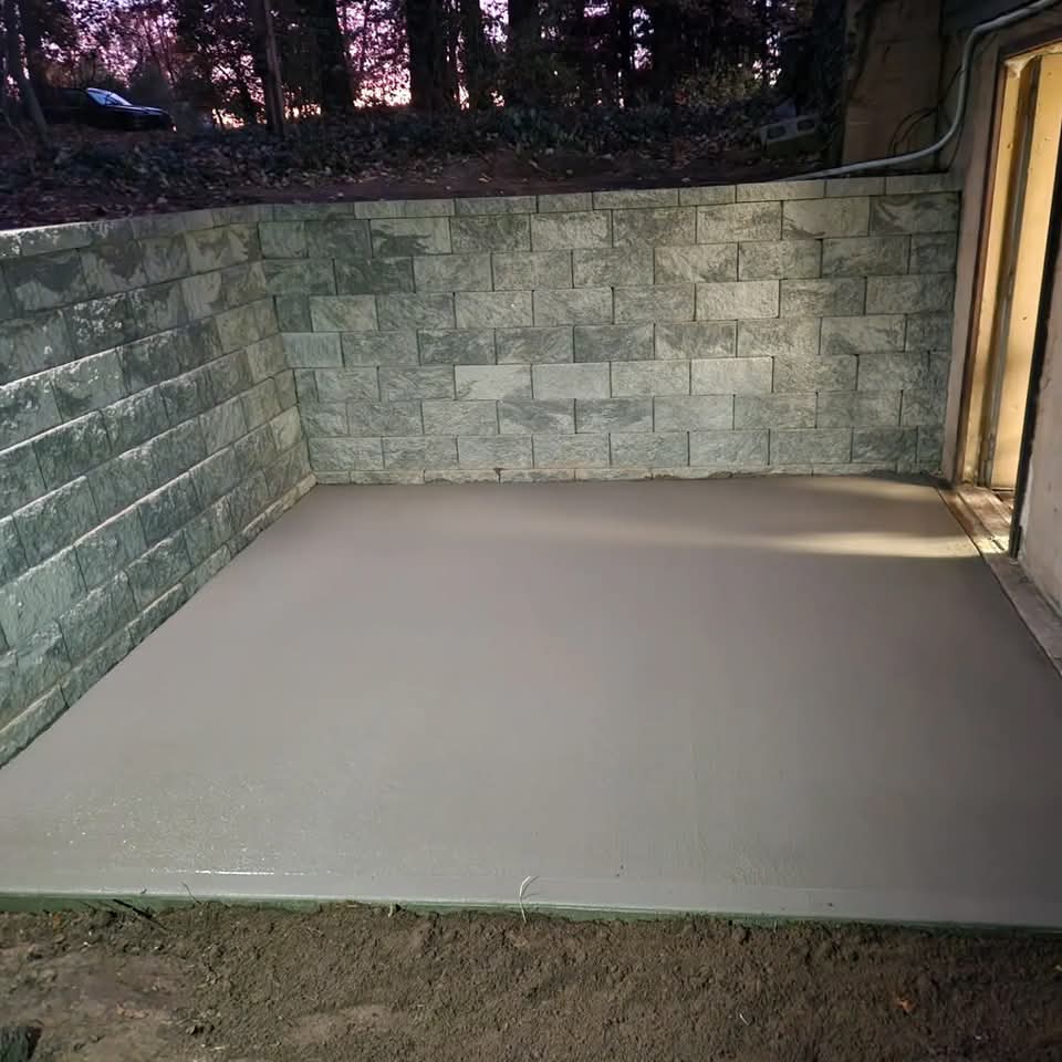 Freshly poured concrete pad enclosed by a block retaining wall on three sides with a wooded backdrop at dusk