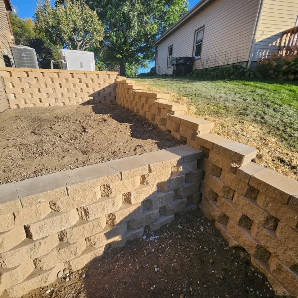 Two-tier tan block retaining wall with integrated steps on a sloped backyard with AC unit and green lawn