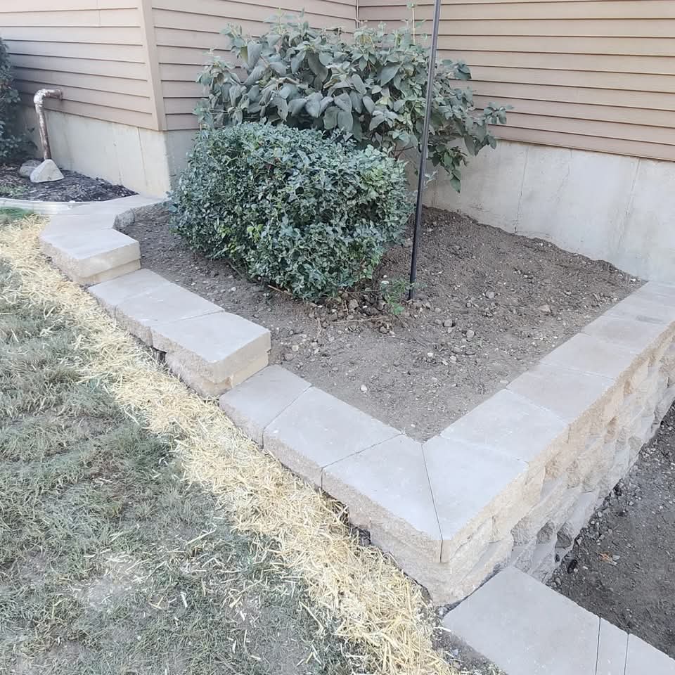Low block retaining wall planter bed with boxwood shrubs and a young tree next to a house foundation with straw-covered seed