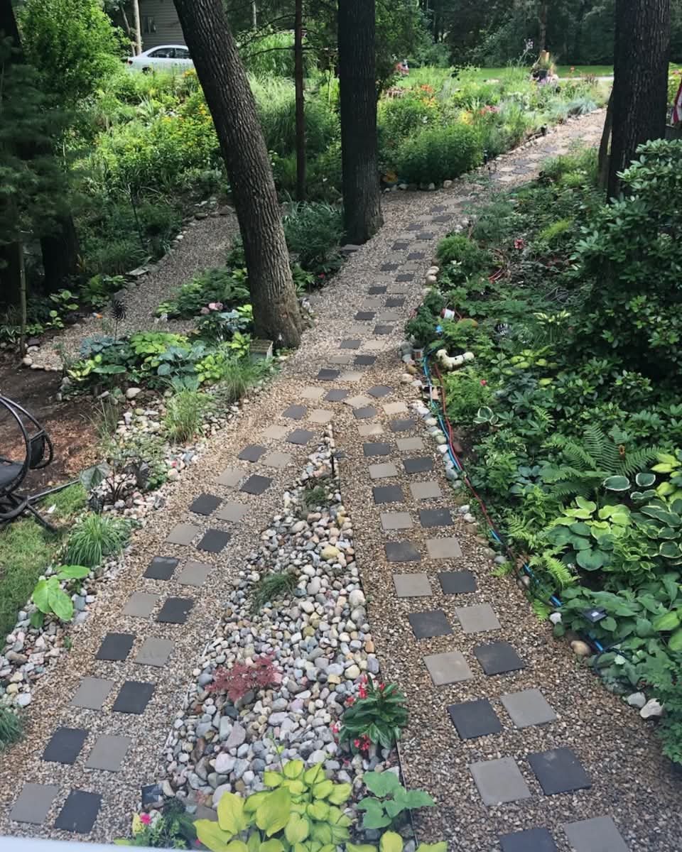 Gravel stepping-stone path through a lush shade garden with hostas, ferns, river rock, and mature trees