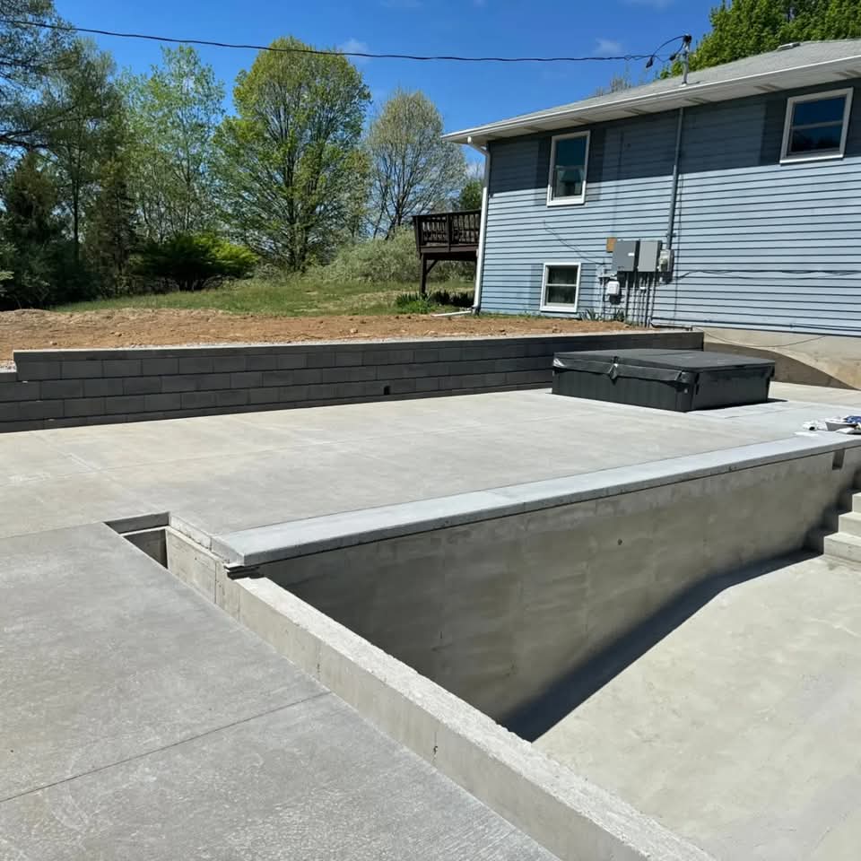 Concrete pool deck and in-ground pool shell with a dark block retaining wall behind a blue house on a graded hillside