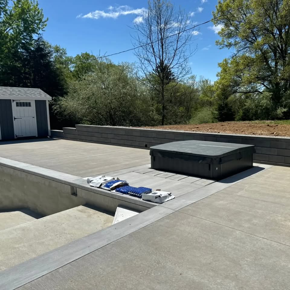 Wide view of concrete pool deck with a covered hot tub, in-ground pool, dark block retaining wall, and a gray storage shed