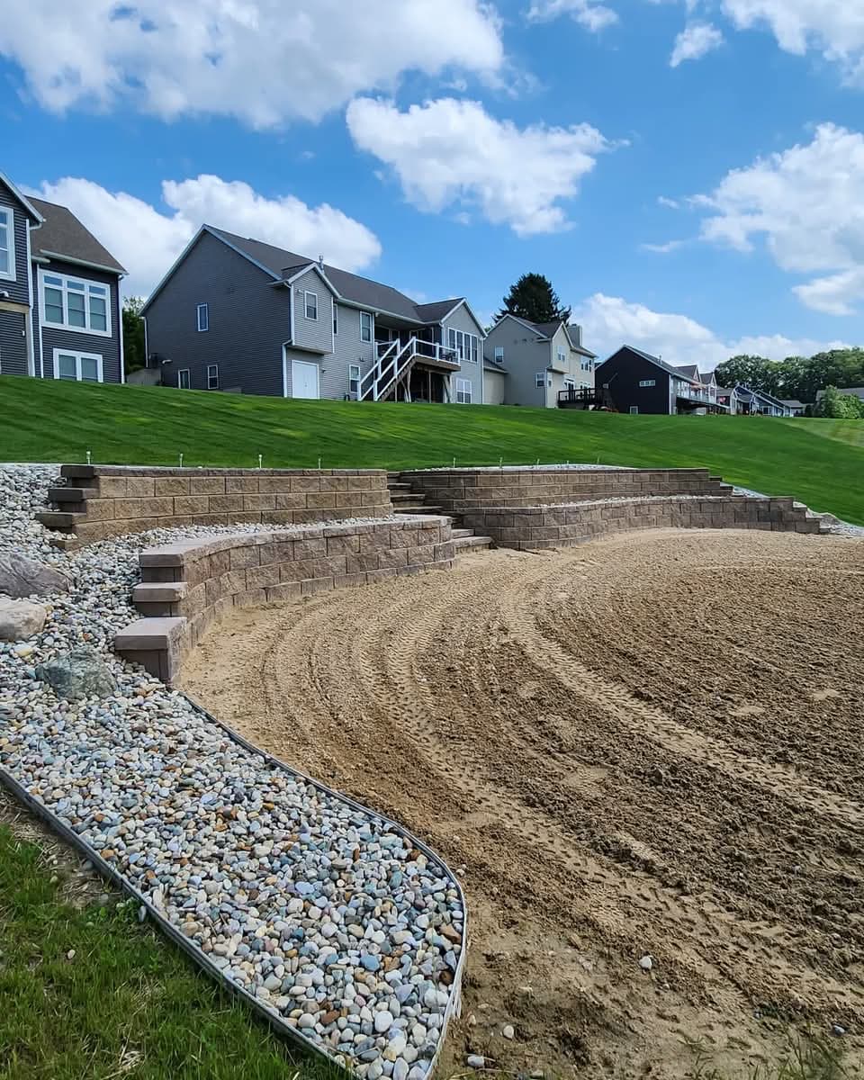 Curved multi-tier retaining wall with river rock edging and graded soil, houses on a green hillside behind