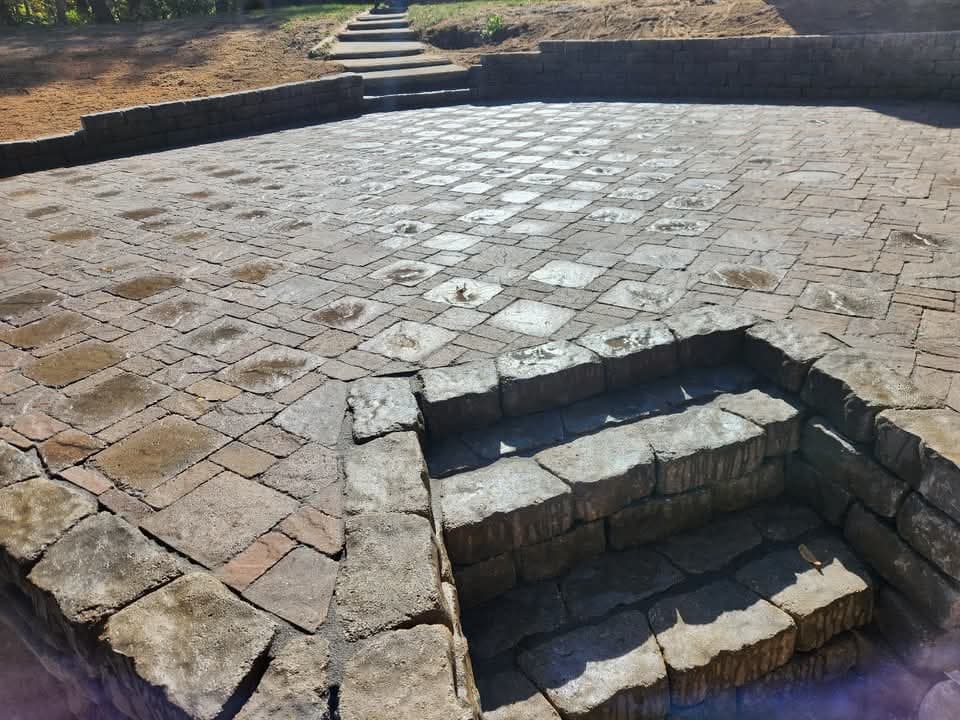 Large curved multi-tone paver patio with a concrete border extending beneath a second-floor deck of a cream-sided house, a natural boulder retaining wall with mulch on the far side, and a green lawn edge