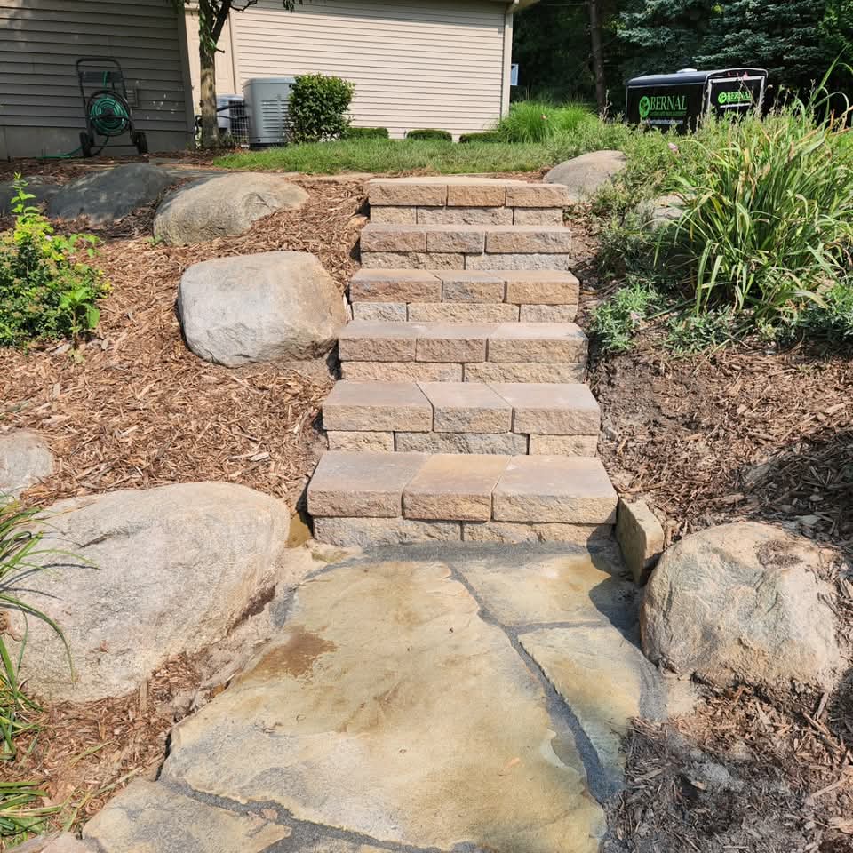 Tan paver stairs descending to flagstone through a freshly mulched bed with boulders and a Bernal yard sign