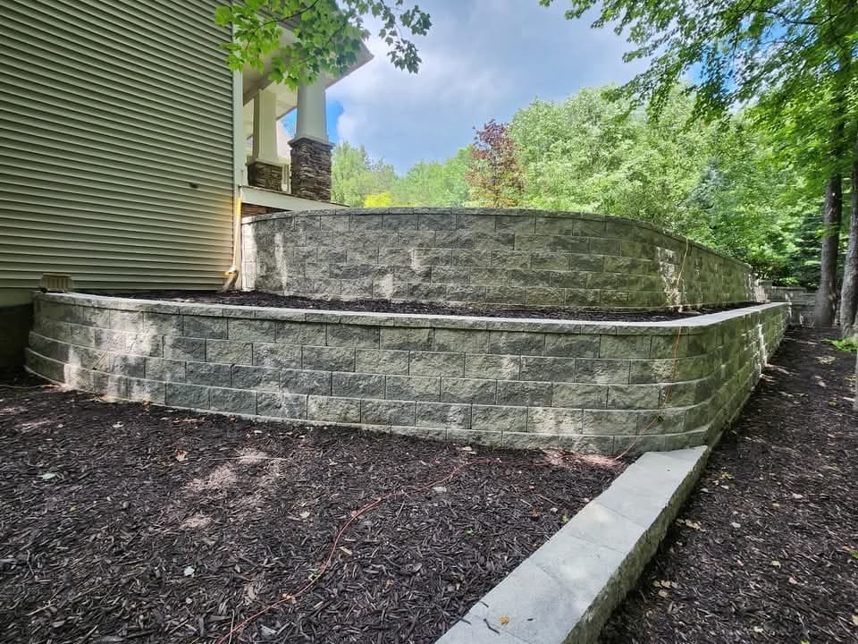 Paver patio surface with a built-in block fire pit in the foreground, block steps leading up to a retaining wall, bare dirt grading on the hillside above, and a wooded backdrop