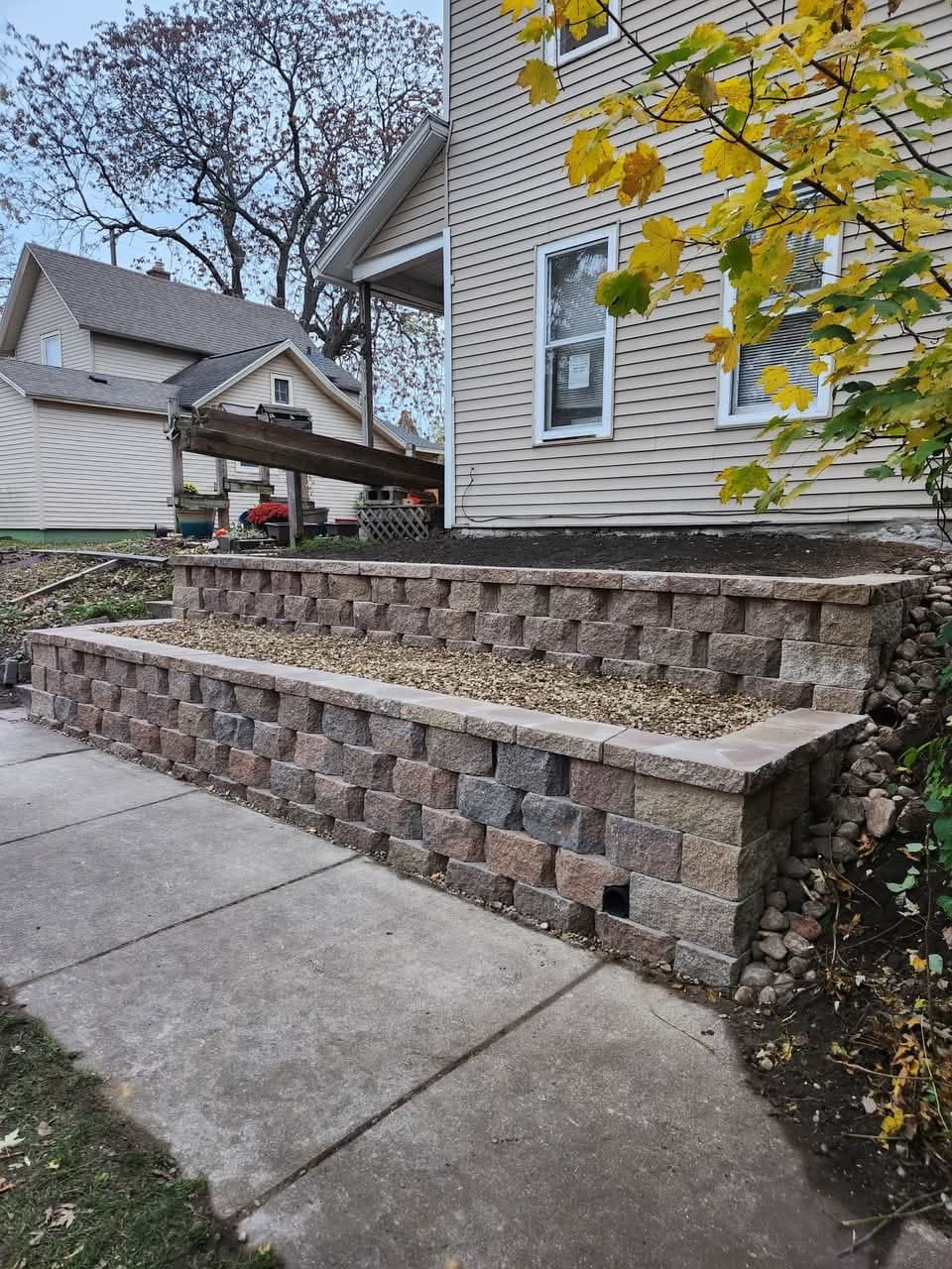 Two-tier block retaining wall with gravel-filled built-in planter next to a concrete sidewalk in autumn