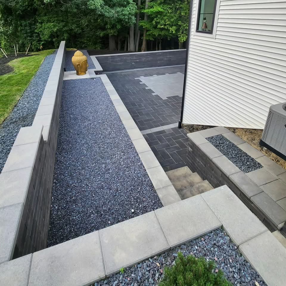Elevated view of dark block retaining walls with gravel beds, a dark paver patio with geometric light-color inlay, a gold statue on a block pedestal, and block steps beside a white house