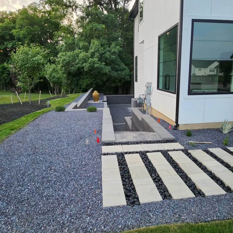 Large-format paver stepping stones with river rock between in a gravel bed, dark block retaining wall with integrated steps, sunken paver patio area, and a gold statue beside a modern house