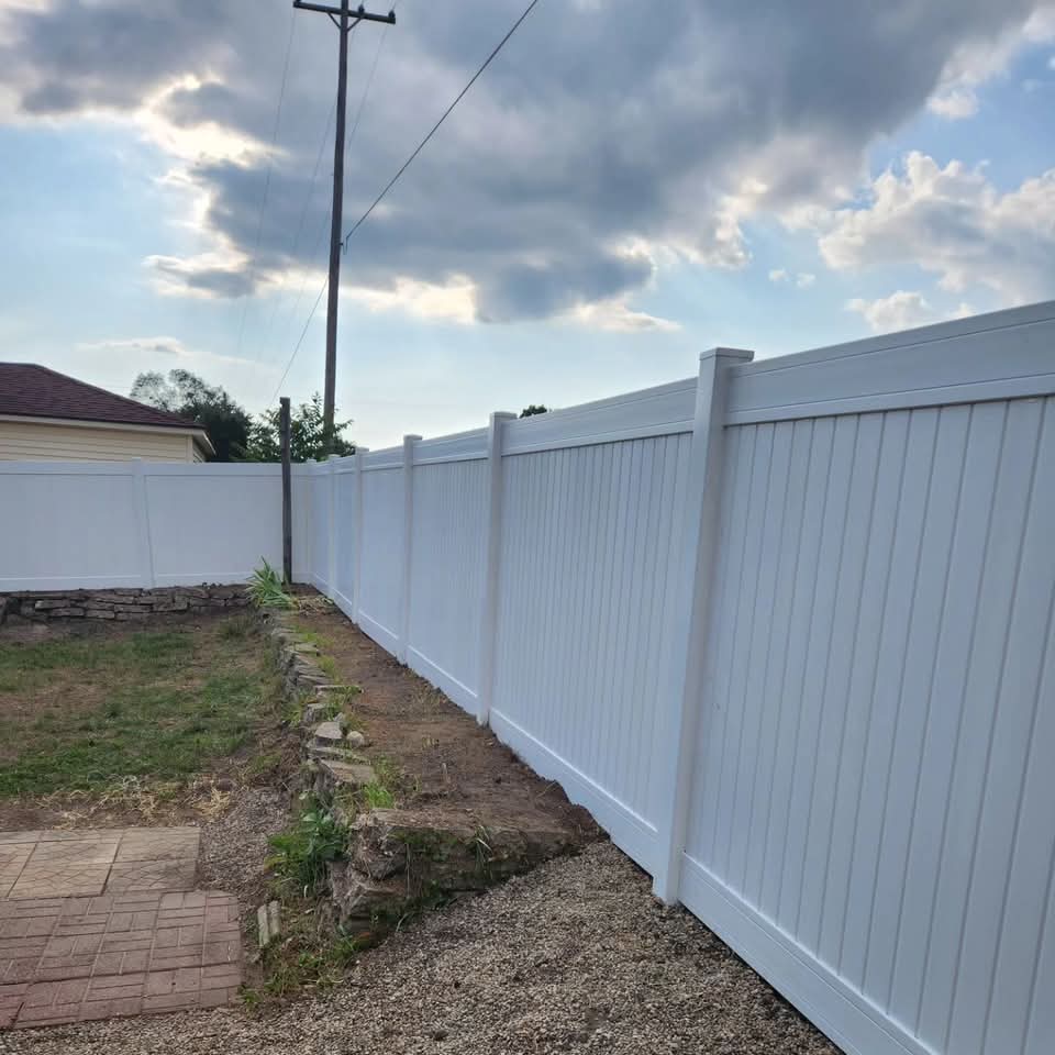 Side of a modern white house with dark block retaining wall, integrated steps, gravel bed, paver patio, trimmed shrubs, and an AC unit with subdivision homes behind