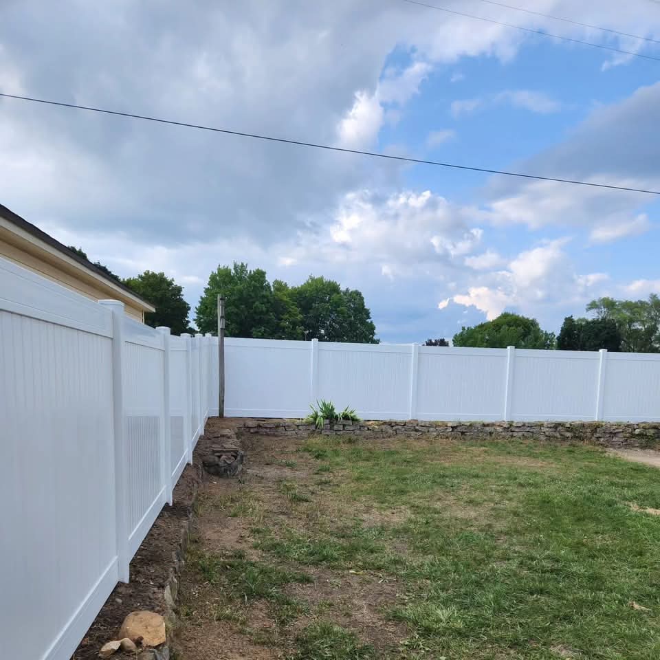 White vinyl privacy fence with a double gate and black hardware next to a red brick house with bare dirt and green lawn in the foreground