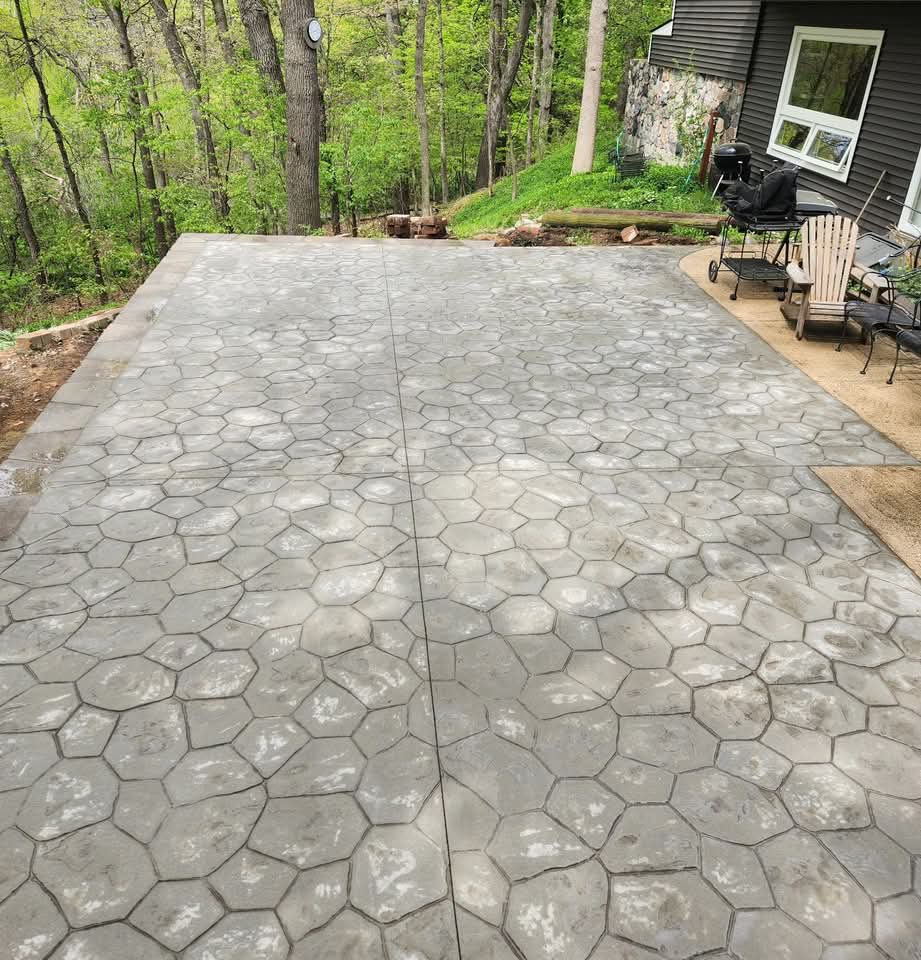 Large stamped concrete patio with an irregular stone pattern behind a dark gray house with stone veneer accents, an Adirondack chair and metal patio furniture, a grill, and a wooded hillside behind