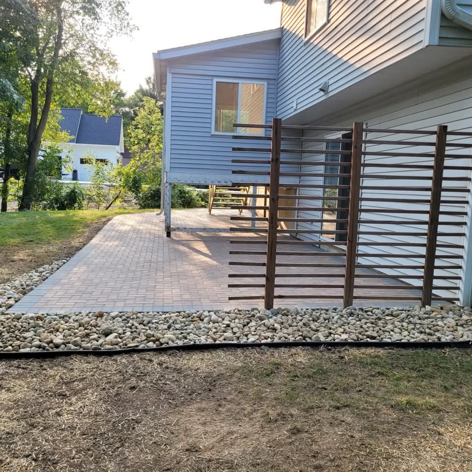 Red-brown paver patio beneath a blue-sided two-story house with a horizontal wood slat privacy screen, river rock border with landscape edging, and straw-covered new seed on the surrounding yard