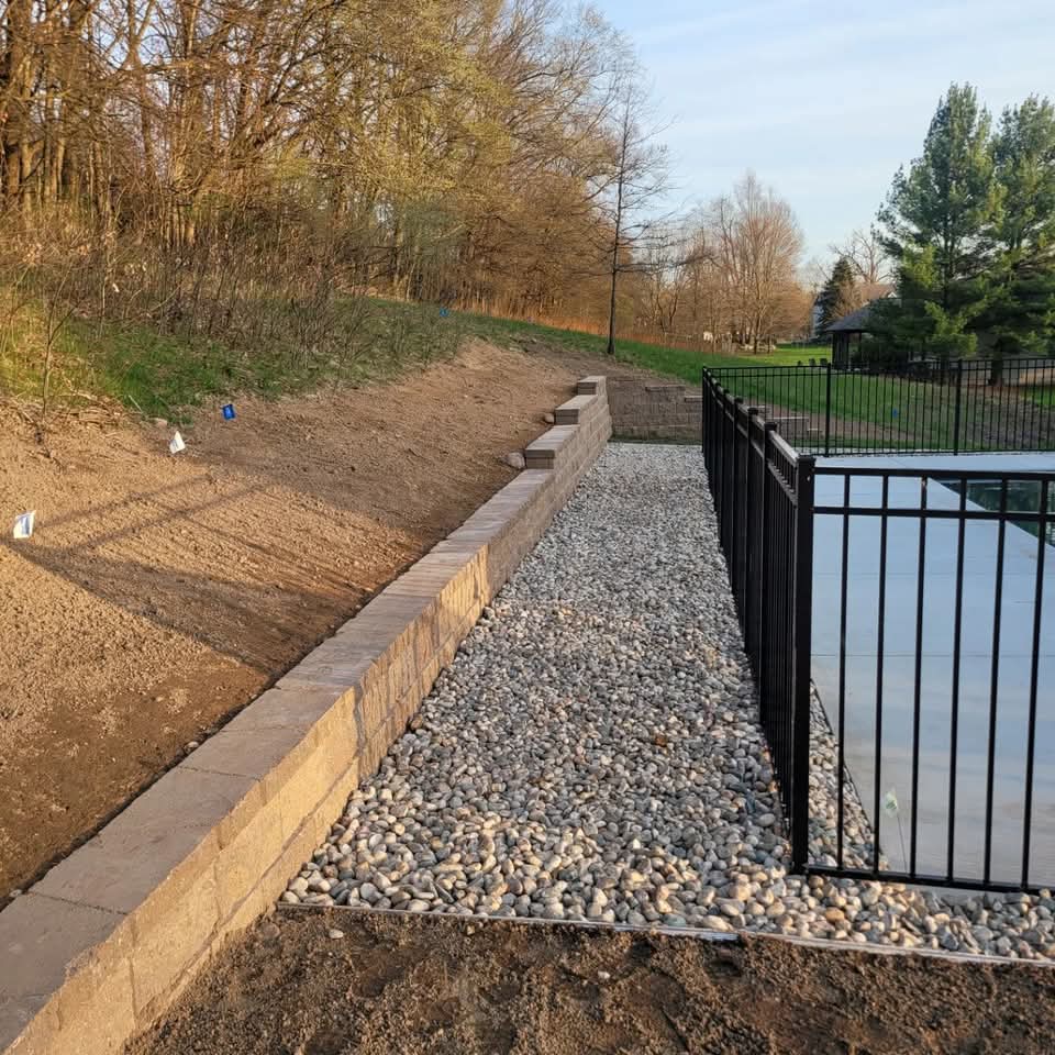 Long tan block retaining wall with cap stones and a river rock strip running alongside a black aluminum pool fence, bare dirt on the uphill side, wooded hillside with early spring trees in the background