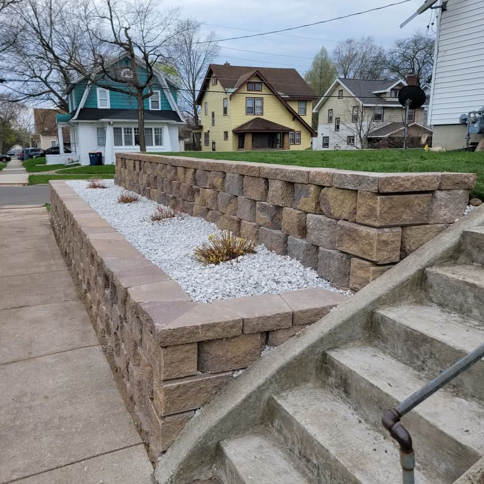 L-shaped brown block retaining wall with white stone planter fill and small plants next to concrete steps with a metal handrail, a teal house and yellow house visible across the street in a residential neighborhood