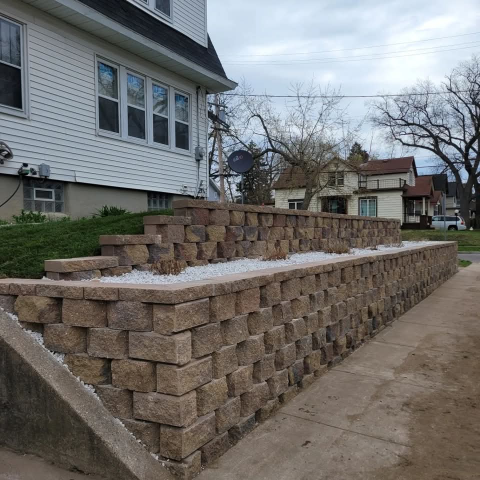 Stepped brown block retaining wall with white stone planter fill along the side of a gray-sided two-story house with a satellite dish, concrete sidewalk below, and residential homes across the street