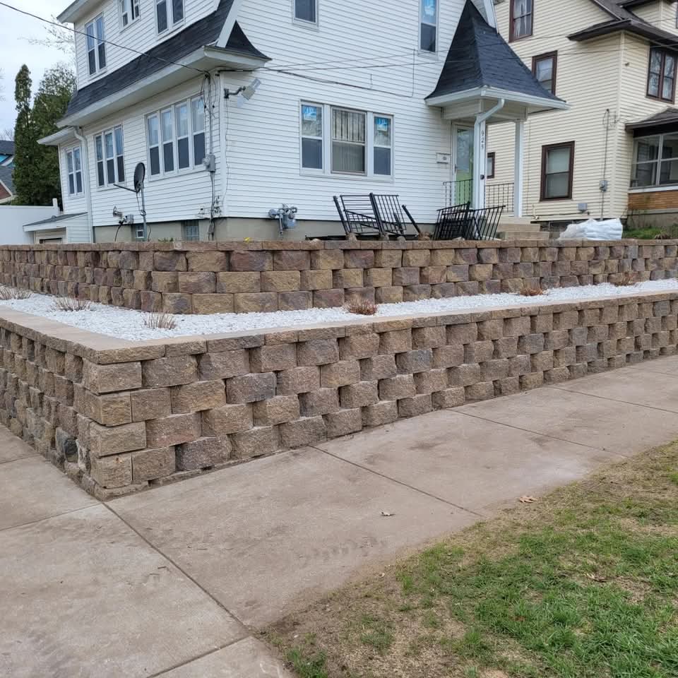 Corner view of a tall brown block retaining wall with white stone planter fill at the top, black rocking chairs on the elevated porch above, concrete sidewalk and green lawn below in a residential neighborhood