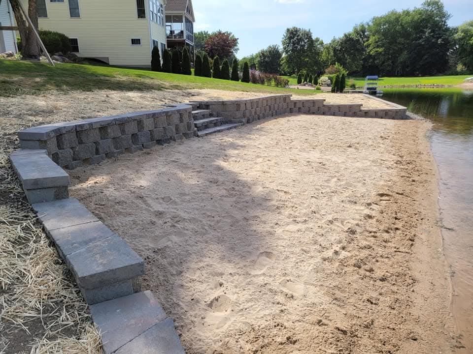 Gray block steps with flat paver treads leading up from a sand beach area to a grassy yard with straw-covered new seed, block retaining wall wing walls on both sides