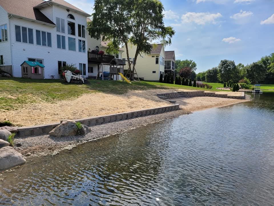 Block retaining wall with cap stones and block steps along a lakefront shoreline, a sand beach area below, a yellow two-story house and neighboring homes on the grassy slope above, and a boat dock on the water