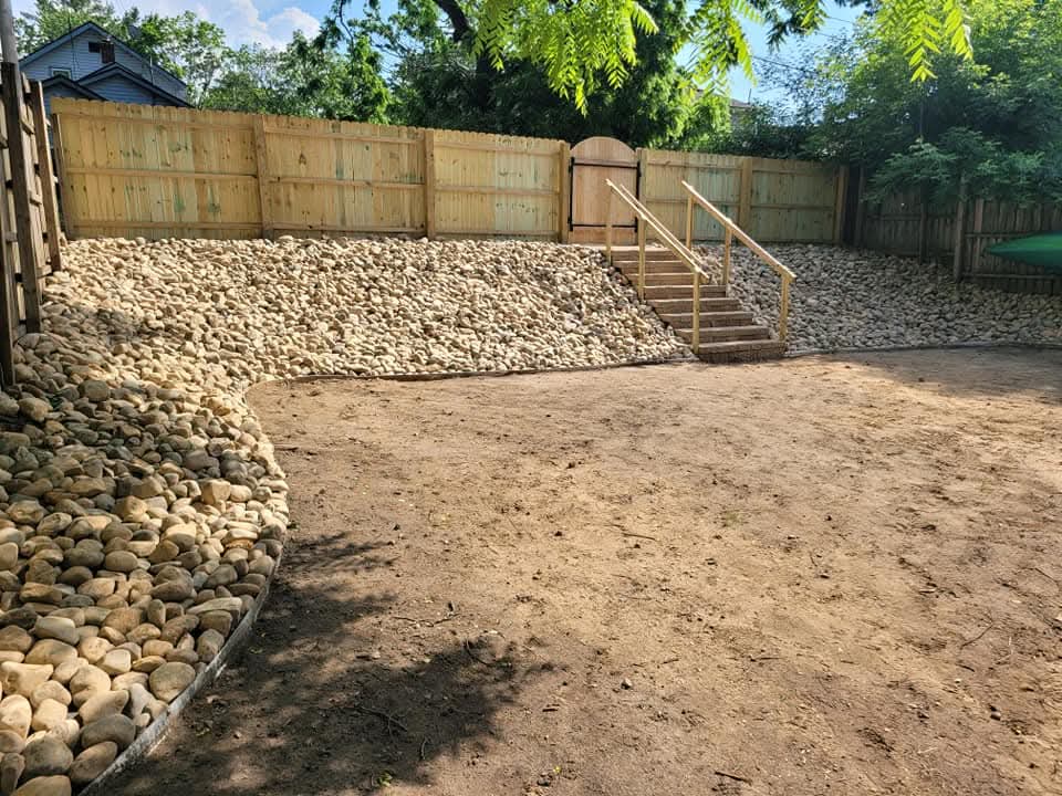 New pressure-treated wood privacy fence running along a residential property line with freshly graded dark topsoil in the foreground, a red-sided house and gray house visible beyond the fence, mature trees overhead