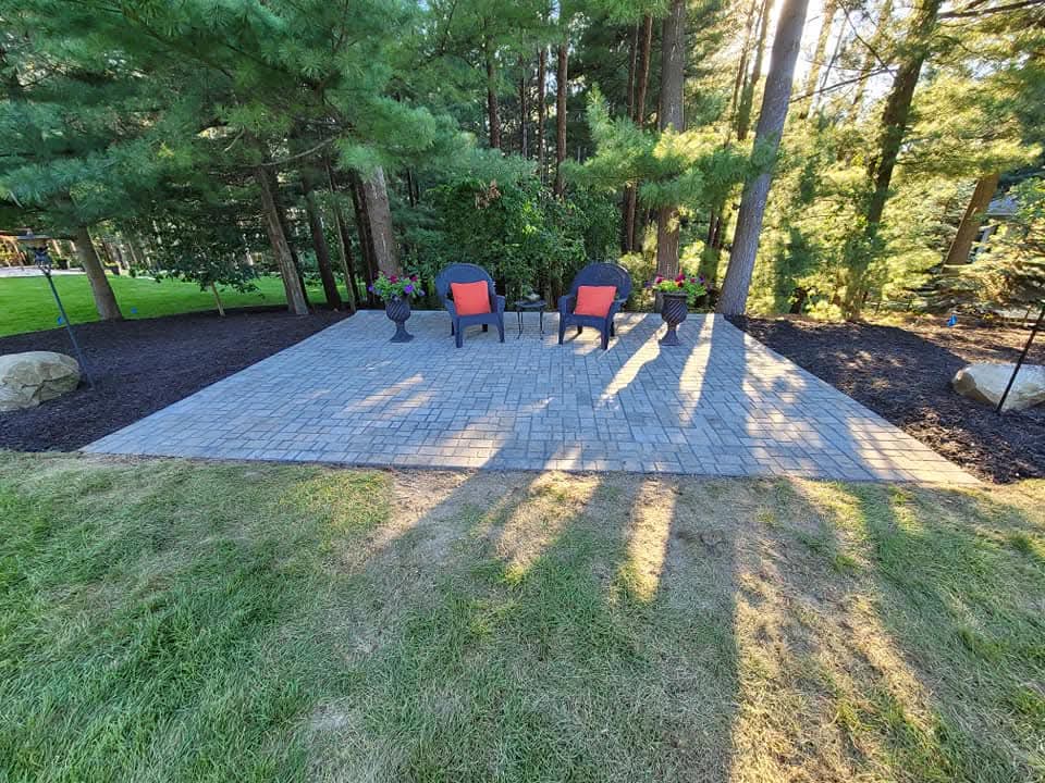 Rectangular gray paver patio with two dark wicker chairs with red cushions and a small side table, bordered by dark mulch beds and natural boulders, surrounded by tall pine trees with dappled sunlight