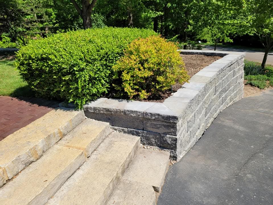 Gray block retaining wall with cap stones along a curved asphalt driveway viewed from stone steps, rounded green and gold shrubs in the mulch bed, brick paver walkway on the left, mature trees in the background