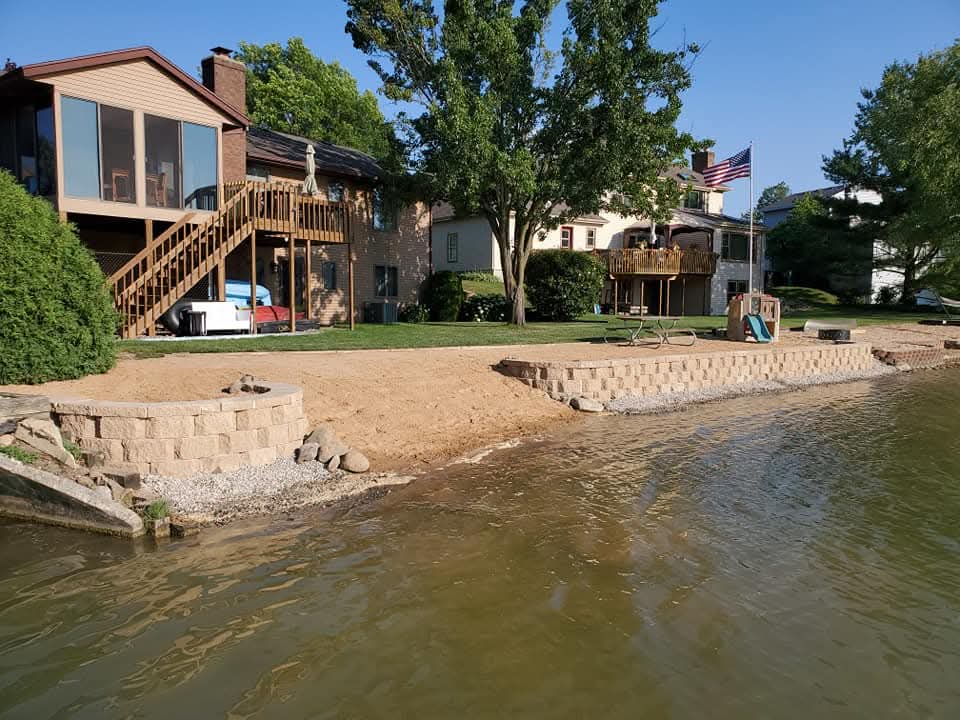 Lakefront property with tan block retaining walls and a round block fire pit on a sand beach area, a brown brick house with wood stairs and a screened porch on the left, a yellow house with an American flag on the right, water in the foreground