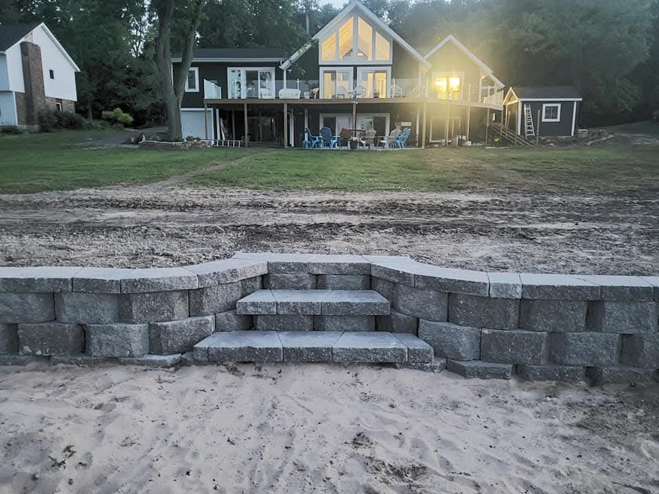 Gray block retaining wall with cap stones and center block steps on a lakefront beach, sand in the foreground, bare dirt grading above the wall, a dark-sided A-frame house with a covered porch and Adirondack chairs on the lawn behind, dusk lighting
