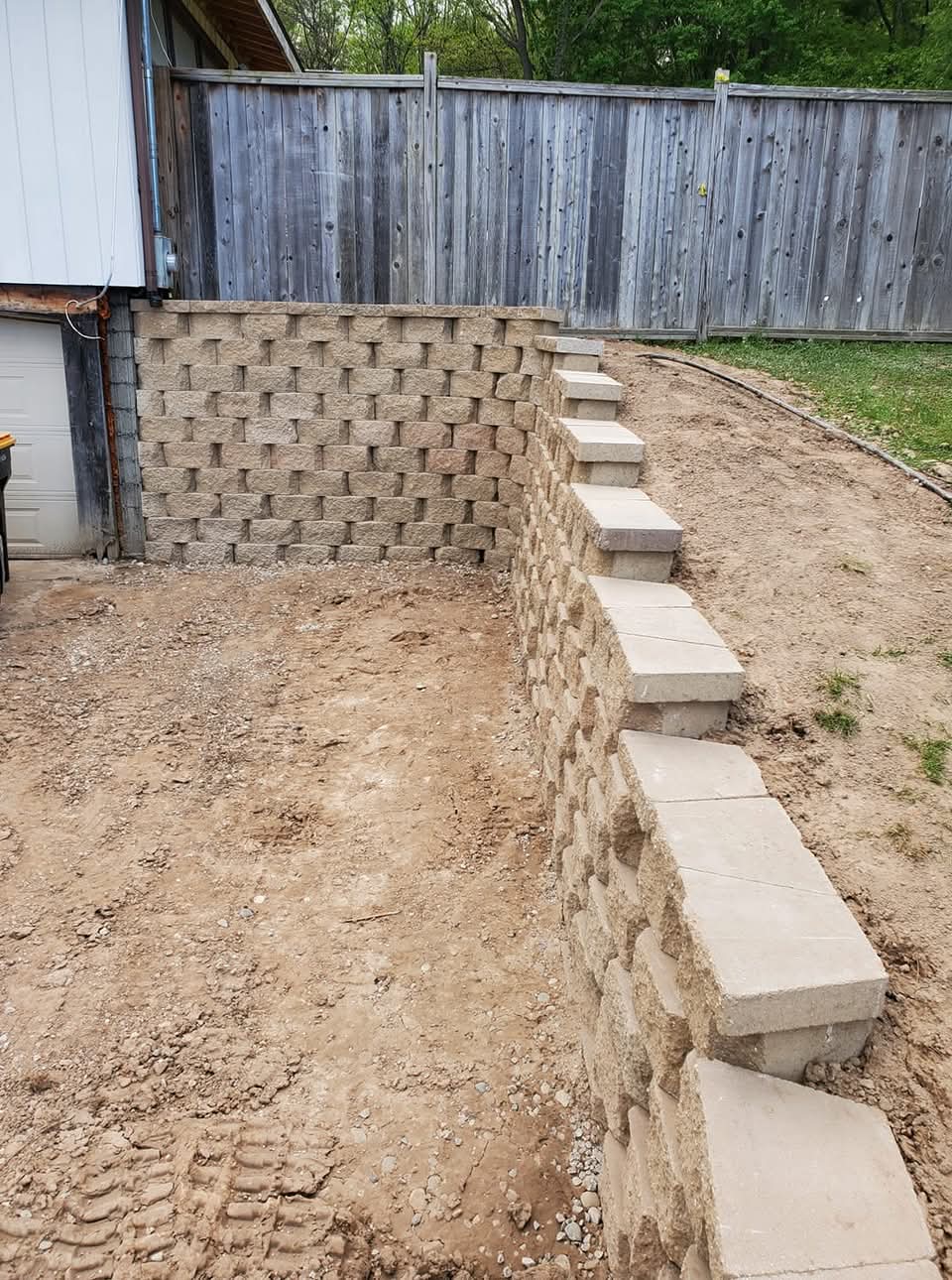 Tumbled block retaining wall with stone steps beside a garage and a wooden privacy fence in background