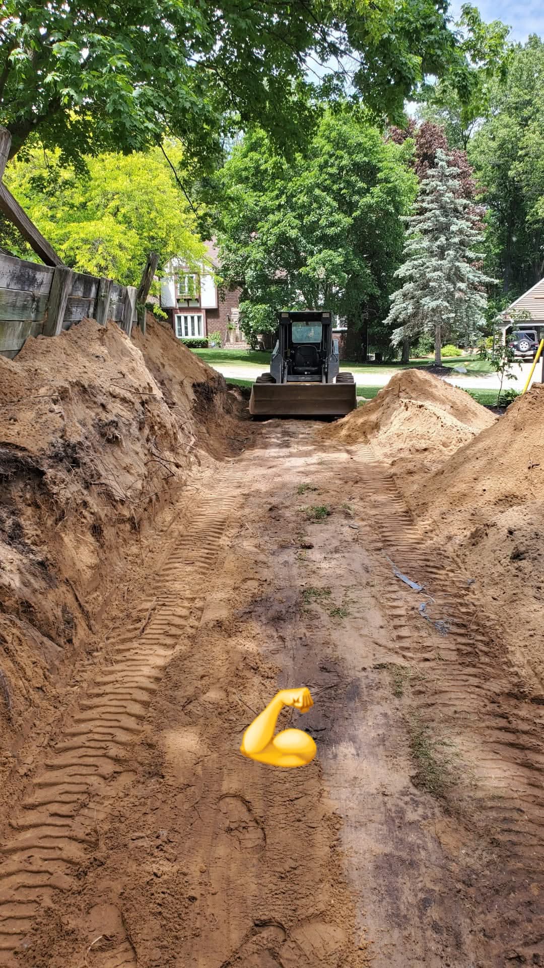 Long deep trench cut across a residential yard with a skid steer at the far end during a drainage install