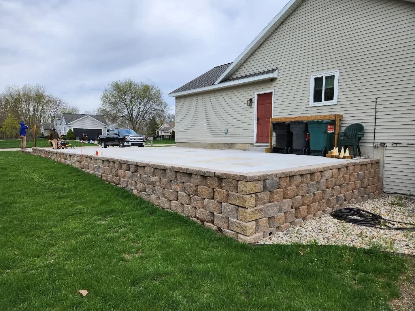 Raised concrete pad on a block retaining wall behind a house with crew workers and green lawn in foreground