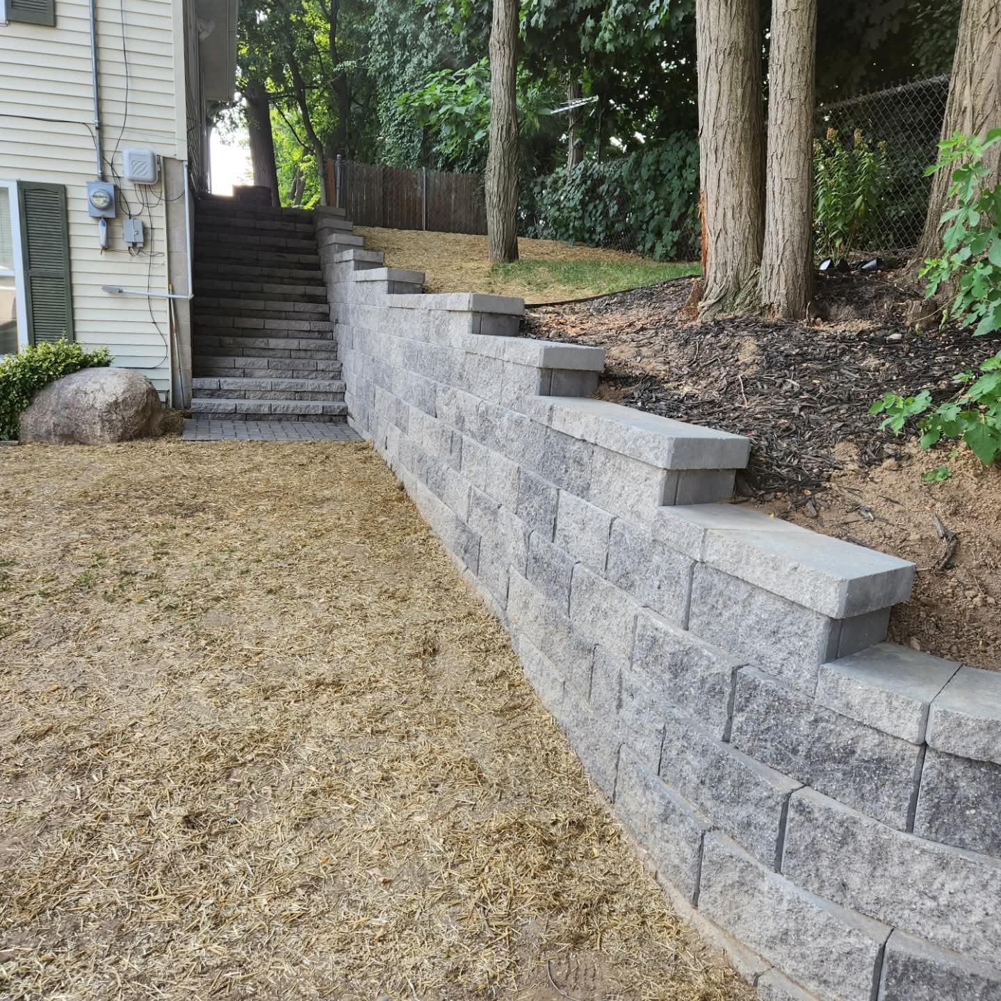 Curved gray block retaining wall with built-in steps alongside a home and a mulched bed on top