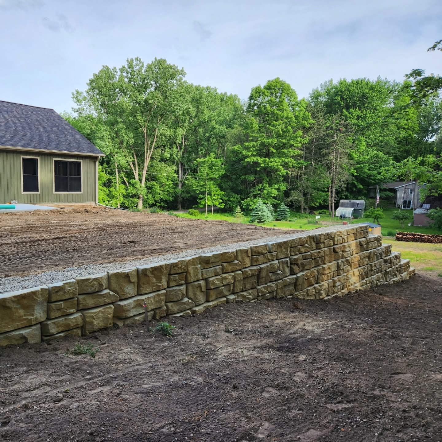 Natural stone retaining wall with gravel base on a graded lot next to a green building with wooded backdrop