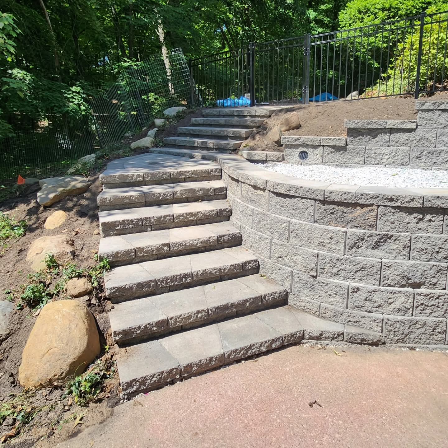 Long block staircase with retaining walls climbing a wooded hillside, curved wall with white gravel and drain grate