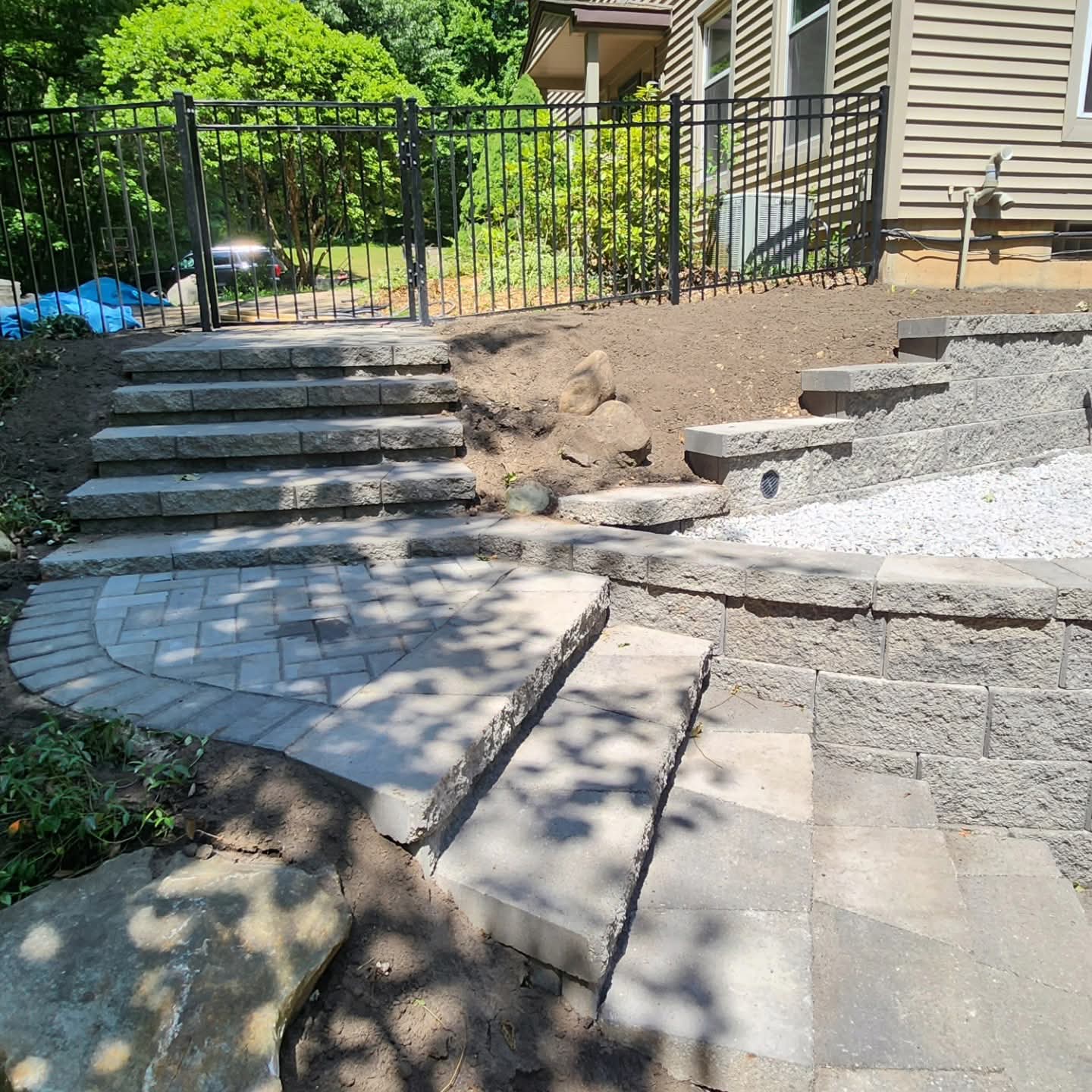 Fan-shaped paver landing at the base of block steps with retaining walls, black metal railing above, and boulders